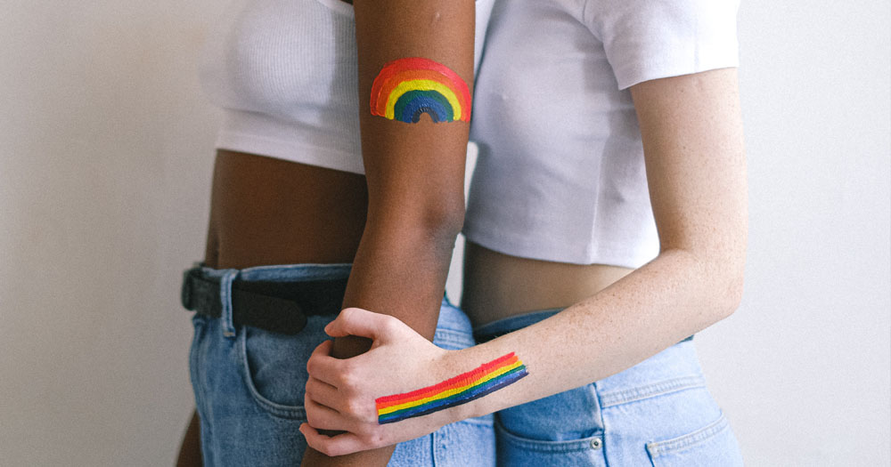 The UK has extended it's pardon of homosexual activity to women and veterans. The image shows two female presenting people standing behind one another. both are wearing jeans and white belly-tops with rainbows painted on their arms.