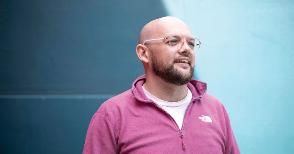 The photo shows online activist Yusuf Murray. He is facing right and has a bald head, glasses and a close groomed beard. He is standing in front of a light blue wall wearing a pink sweater over a white tshirt.