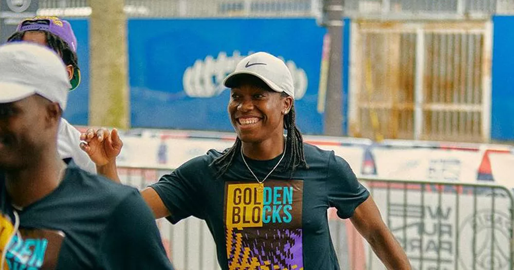 The image shows runner Casterse Menya in a warm-up pose for a run. She has long braided hair and is wearing a grey Nike baseball cap and a black tshirt with the words "Golden Blocks" on it.