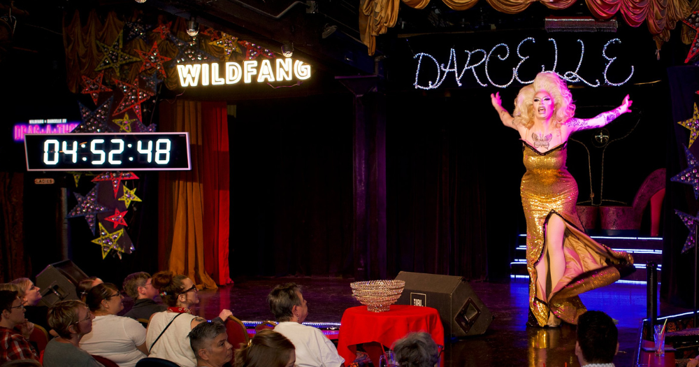 A drag queen in a golden sparkling dress performs on the Darcelle XV stage while breaking the Guinness World Record for Longest Drag Performance to a large crowd.