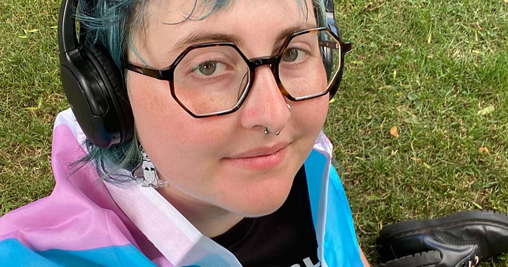 Non-binary person smiling toward camera wearing glasses and trans flag