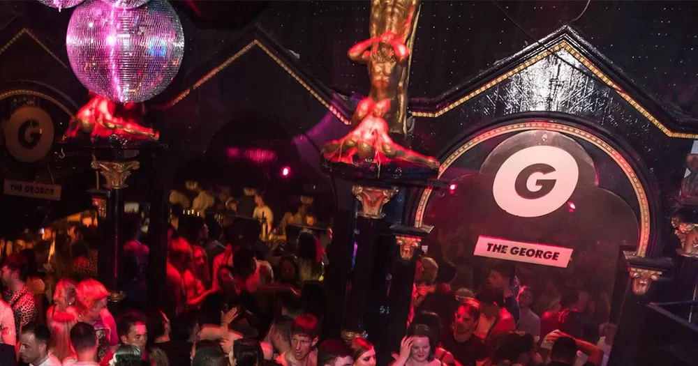 The dance floor at Dublin's most popular queer nightclub, The George, bustles with a large crowd. A disco ball hangs on the left and The George logo is on the wall on the right.