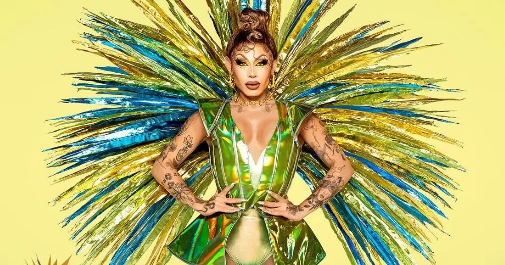 Host of Drag Race Brazil Grag Queen stands fiercley against a yellow background adorned in sparkling yellow and blue feathers while wearing a bright green shining bodysuit.