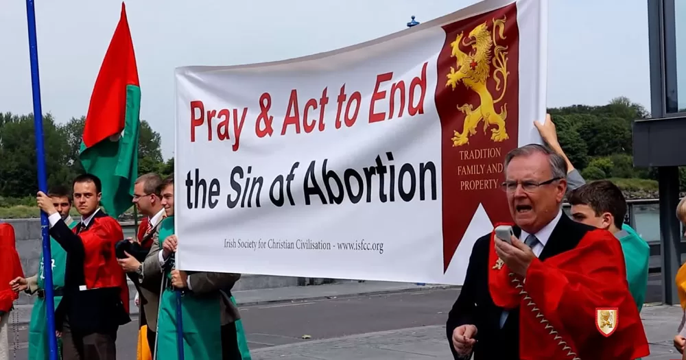 The image shows members of the Irish Society for Christian Civilisation, the orgainsers of 'chivalry camp', at a protest. They are holding up a banner which reads, "Pray & Act to End the Sin of Abortion" with a crest beside it. They are dressed in black suits with red and green sashes over their shoulders. One of the older men at the front is holding a megaphone speaker giving a speech.