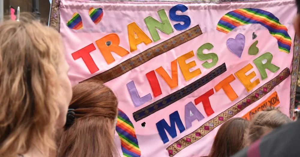 A flag hangs with "trans lives matter" is written in colourful letters with people gathering around in front of it.