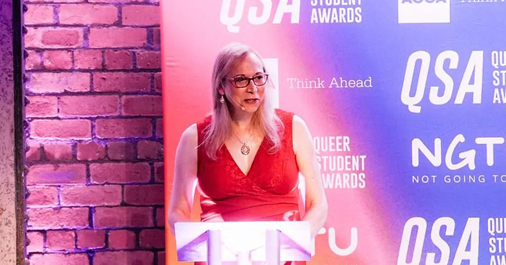 The image shows Katie Neeves who discusses trans inclusion in the workplace in this article. In the image she is standing in front of a podium delivering a speech. She is wearing a red sleeveless dress with a v neck front.