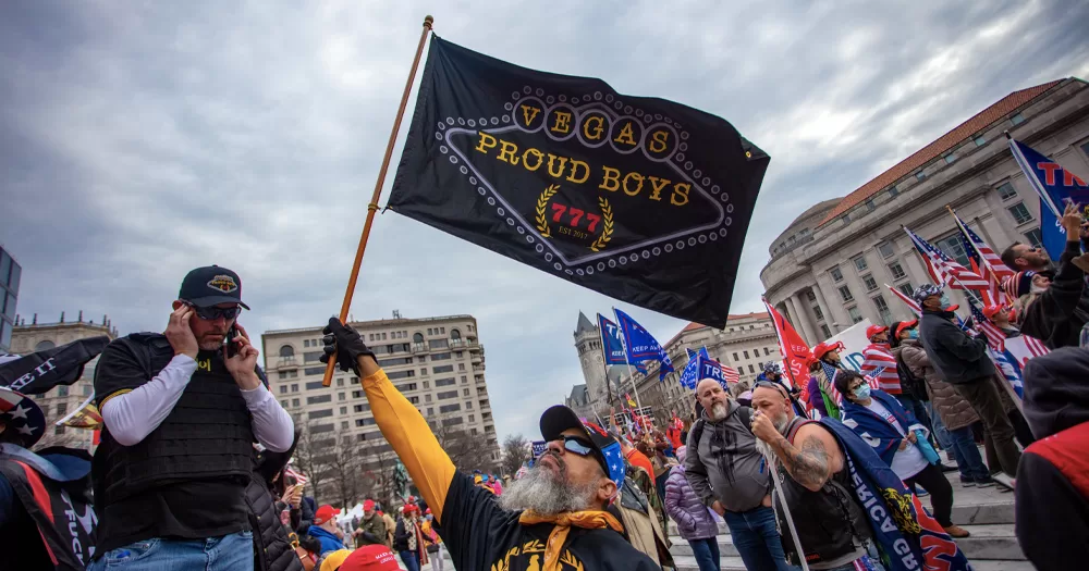 This article is about anti-LGBTQ+ protests in Ireland and the US. In the photo, a man waving a "proud boys" flag with other men in the background with US flags.