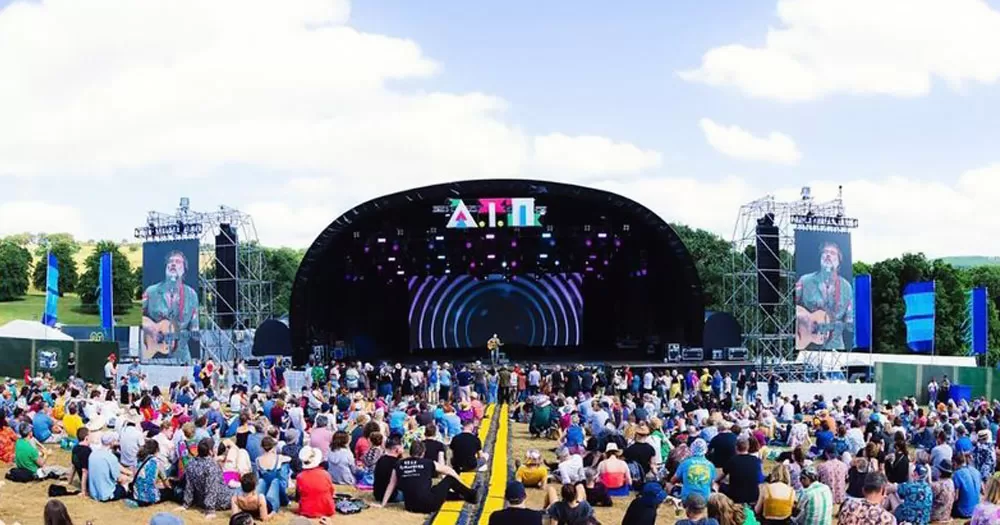 The image is a view of the main stage at the All Together Now festival who have cancelled Osunlade for anti-trans posts. The image shows the stage in the background with people sitting on the ground facing it. It is daytime and there is a yellow and black path running down the centre of the image.