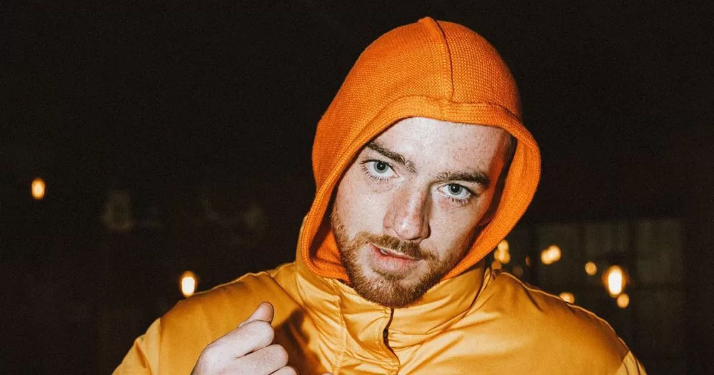 The image shows actor Angus Cloud wearing an orange puffa jacket and orange hoodie. He is wearing the hood up and is biting the side of his lip as he looks directly into the camera.