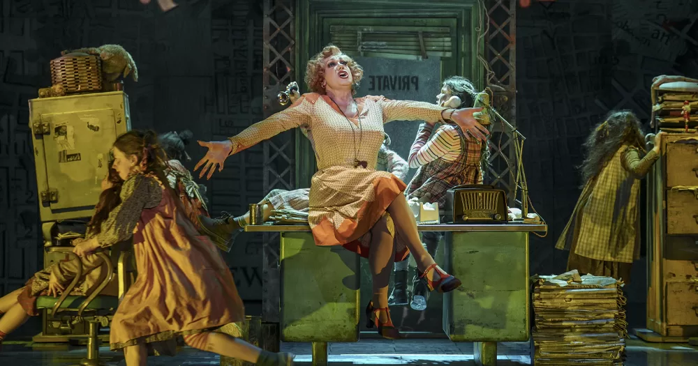 Scene from Annie the Musical, with character Miss Hannigan singing while sitting on a desk and kids working around her.