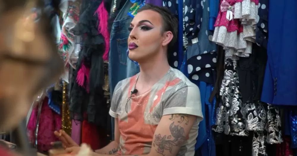 A queer person from Russia with beautiful makeup sits among large rails full of vibrant clothes. This is a scene featured in the moving documentary A Worm in the Heart by Paul Rice.