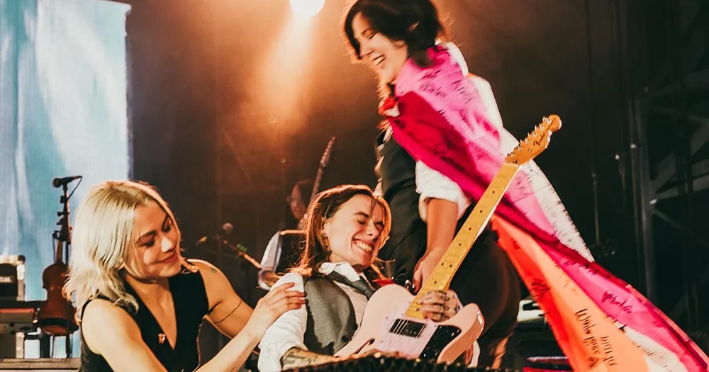 boygenius on stage. Phoebe Bridgers kneels with Julien Baker playing guitar, and Lucy Dacus stands slightly behind them with a lesbian flag draped around her.