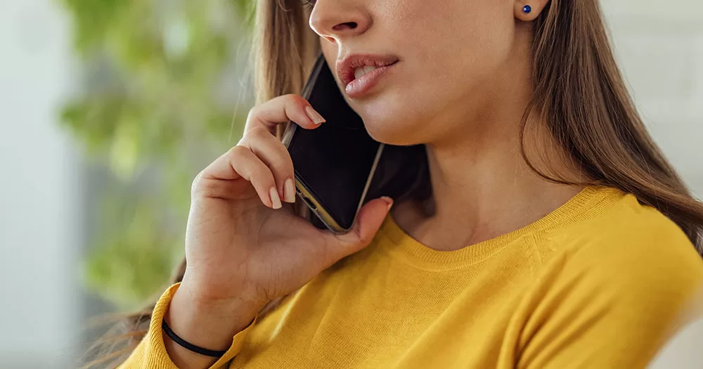 Dublin Lesbian Line and The Switchboard launch 'Women's Night' phone service. The image shows a female presenting person using a mobile phone. She is wearing a yellow jumper and is holding the phone to her ear. She has long brown hair and the image is cropped from her nose to chest.