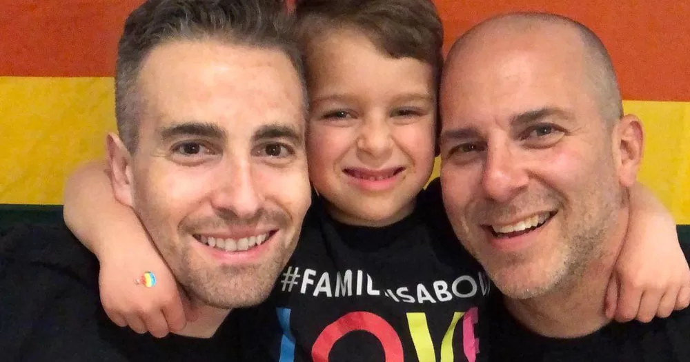 The image shows fathers BJ Barone and Frankie Nelson who won a case against Fratelli d'Italia for the use of their photo. In the image, the two men are standing in front of a Rainbow coloured background, being hugged by their son Milo.