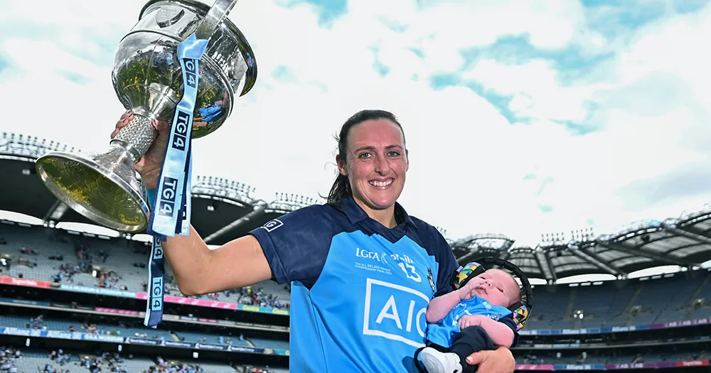 Hannah Tyrrell holding the All-Ireland trophy in one hand and her newborn baby in the other. Hannah is in Croke Park wearing a blue Dublin jersey.