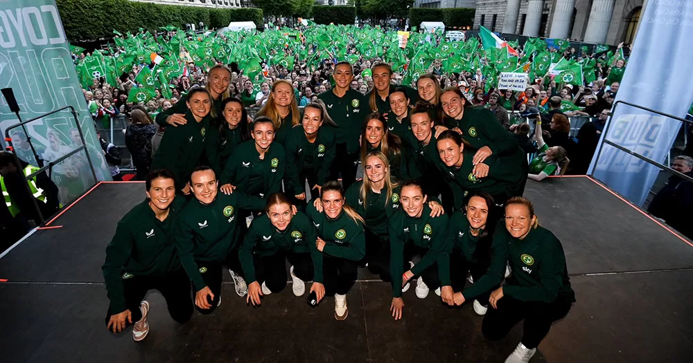 The Ireland women's football team posing on stage with a crowd of fans at their homecoming event.