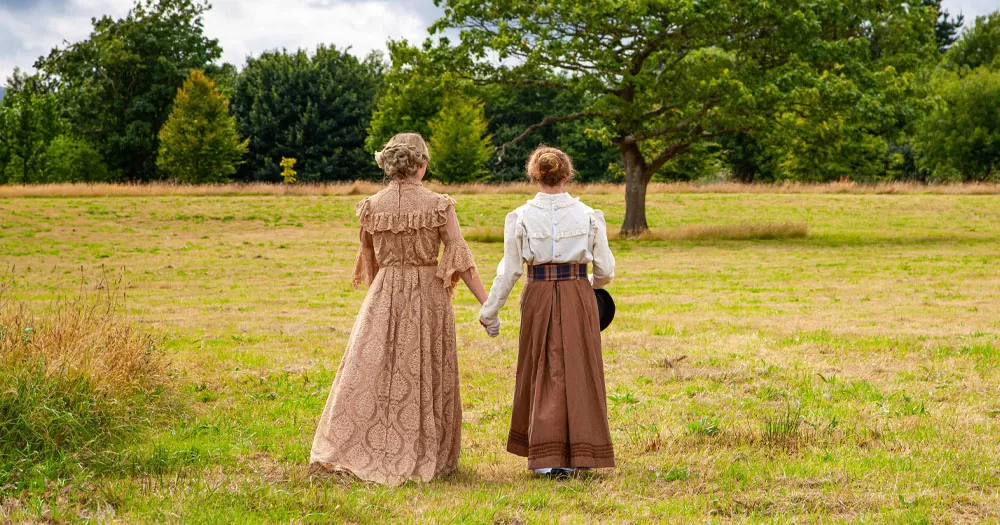 The image shows a promotional shot for the new TG4 documentary of two women posing as the Ladies of Llangollen. The two people are walking in a rural setting holding hands. They are walking away from the camera and are wearing period costumes. The one of the left is wearing aa long tan coloured dress. The one on the right has a long brown skirt with a frilly white blouse. She is also carrying a dark brown hat.