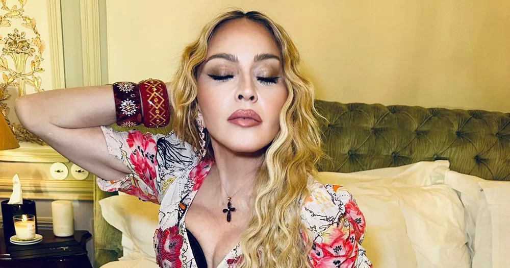 Madonna posing with her eyes closed, sitting on a bed. She wear a patterned dress and a cross necklace.