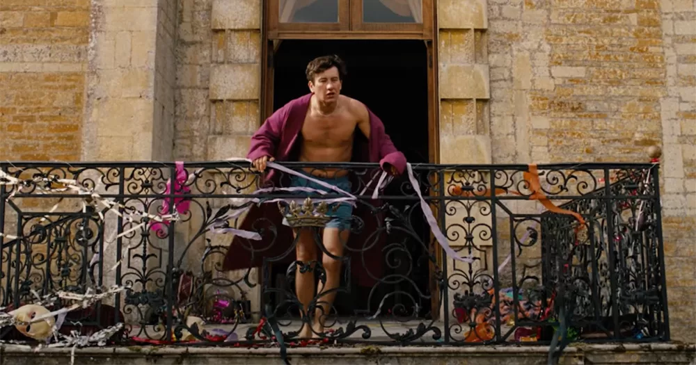 Barry Keoghan in the Saltburn trailer. Keoghan is standing on a balcony in shorts and a robe, and his bare chest is visible.