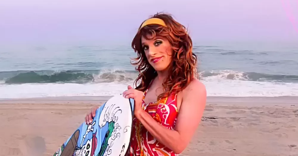 The image features Shirley Temple Bar who has been interviewed in a new podcast. In the photo, the drag queen is standing on a beach holding a surfboard. She has a yellow hairband in her red wig and is wearing a sleeveless red summer dress.