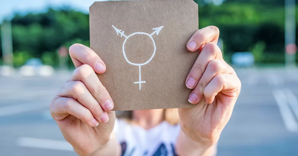 This article is about a trans person coming out in the workplace. In the photo, the hands of a person holding up a sign with a trans symbol.