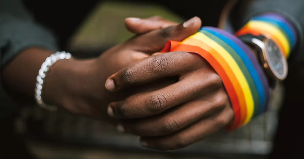 This article is about a man in Uganda being charged with aggravated homosexuality. The image is a close up of hands clasped together with a rainbow band wrapped around one hand and arm.