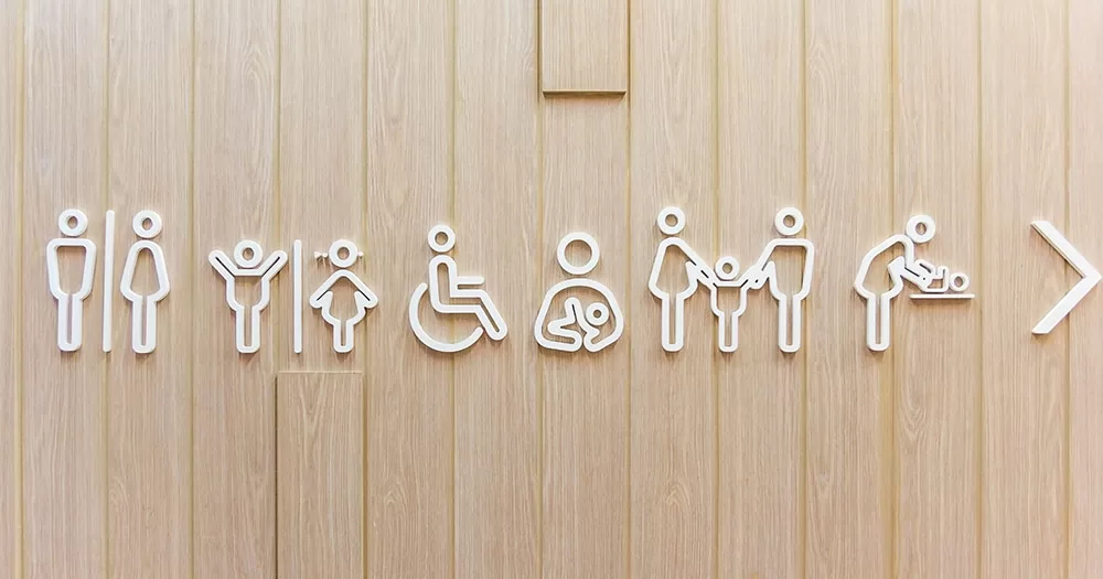 The UK Government are implementing new policies on the the gendered toilets. The image shows a series of cutout graphics representing toilets and facilities mounted on a wooden wall. The graphics include male and female, disabled, parent nursing a baby, parents holding a child's hand, and someone changing a baby.