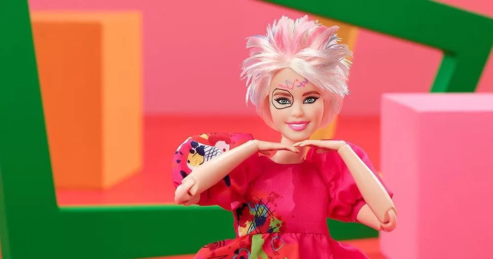 The image shows the new Weird Barbie doll. The doll has short messy pink and blue hair and has colourful markings on her face. She has both her hands positioned under her head as if supporting her chin. She is sitting in front of pink, orange and green geometric shapes.