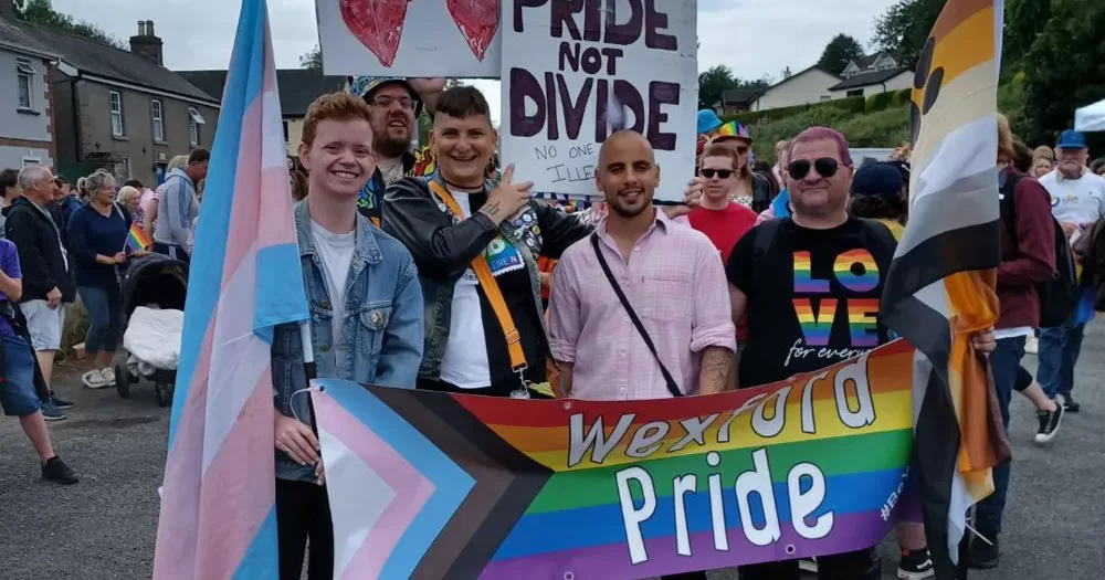 The image shows members of Wexford Pride standing behind a banner at one of the festival events. The banner is painted in the Progressive Pride flag colours with the words "Wexford Pride" in a white bubble font over the rainbow. There are 5 people standing in the picture. The one on the left is holding a trans Pride flag and the person on the right is holding a Bear Pride flag.