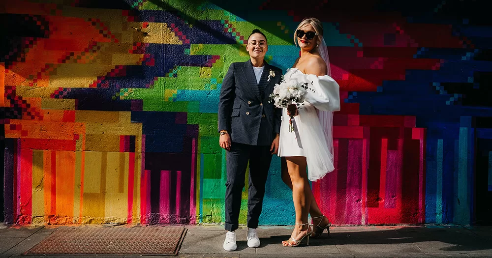A couple, one of which is in a Women's Made To Measure Suit, pose in wedding outfits in front of a colourfully painted wall.