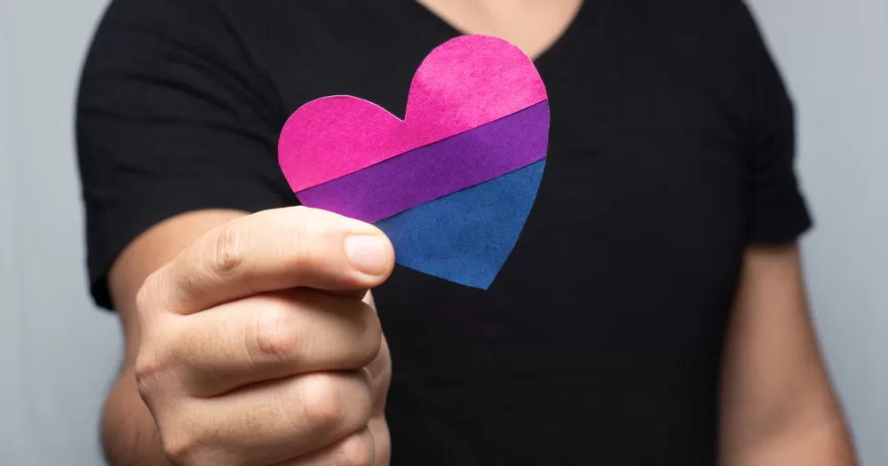 This article is about being a bisexual ally. In the photo, a person holding a heart-shaped bi flag.