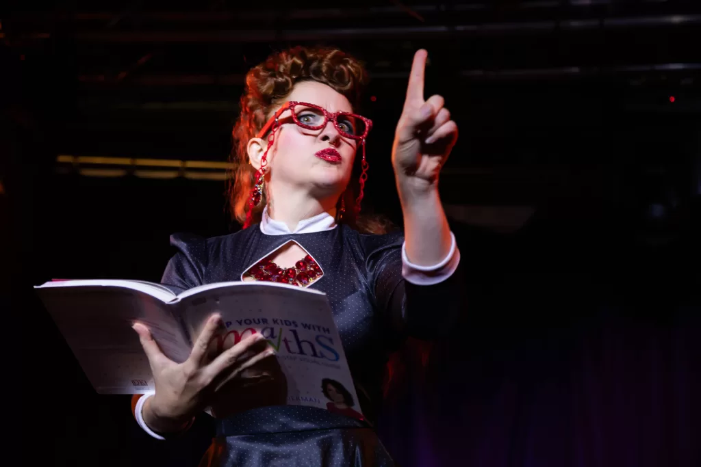 Photo from the Candlelight Cabaret featuring a performer dressed as a teacher holding a book while on stage.