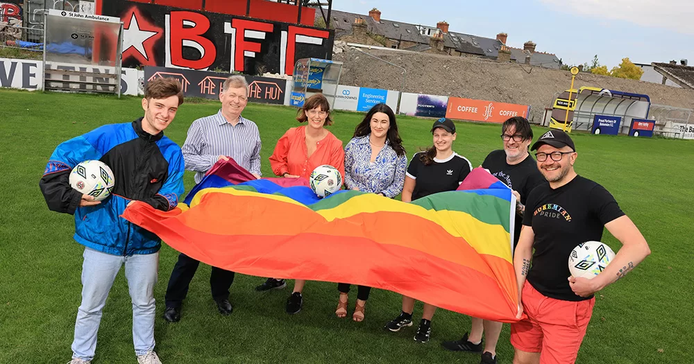 Members of Bohemian FC smiling on the pitch holding an LGBTQ+ Pride flag and footballs.