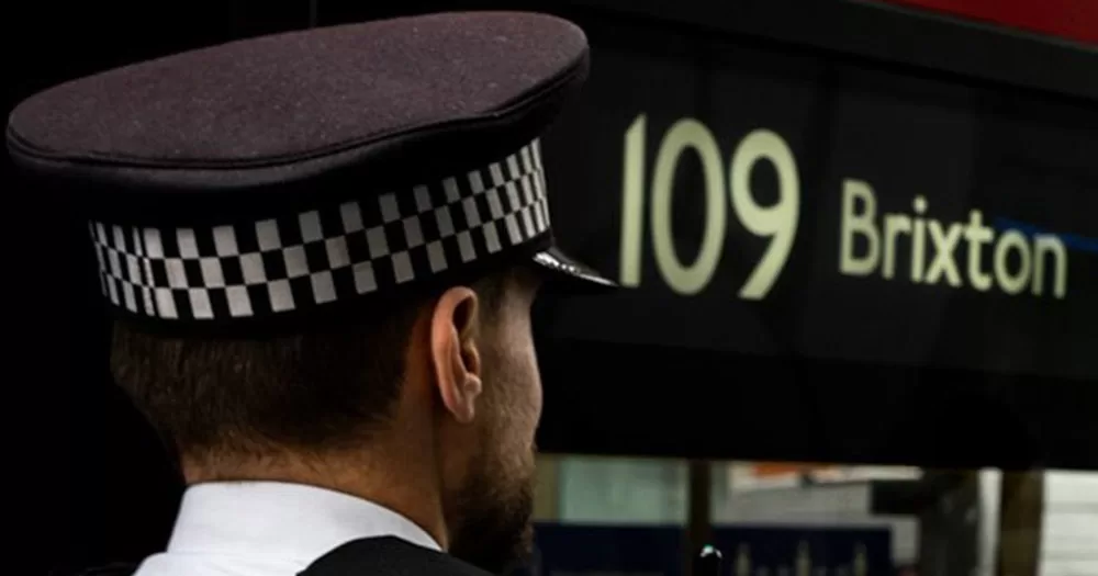 This story is about a man being charged after attacking a gay couple at UK Black Pride. The image shows a close up of the back of a police officer's head, with the sign "109 Brixton" in the background. The police officer is wearing a black uniform cap with checkered print around it.