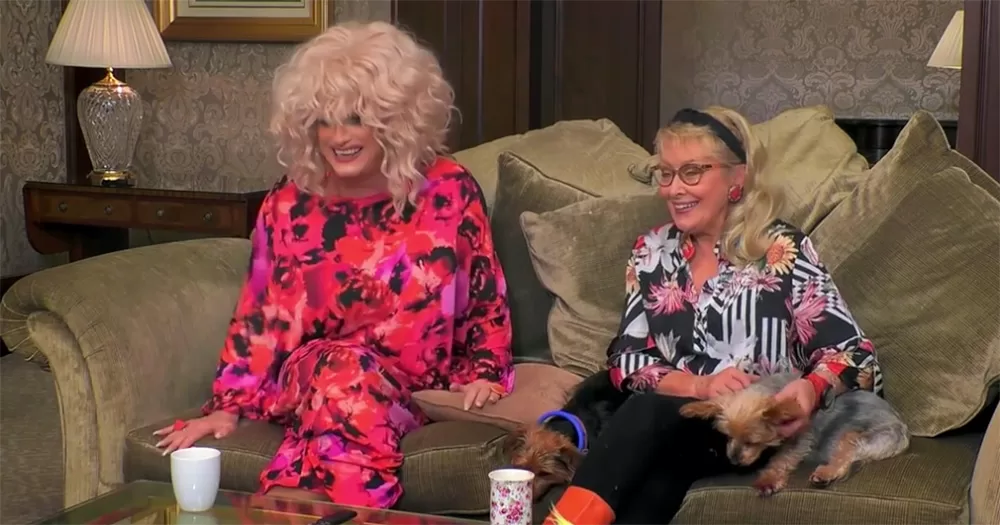 Panti Bliss and Twink in Celebrity Gogglebox Ireland. The pair sit on a beige couch with Twink's small dog. Panti wears a pink patterned dress and blond curly wig, and Twink wears a patterned shirt and black trousers.