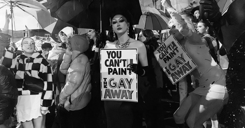 A drag queen at the Chambers Bar protest holds a sign reading "You can't paint the gay away".