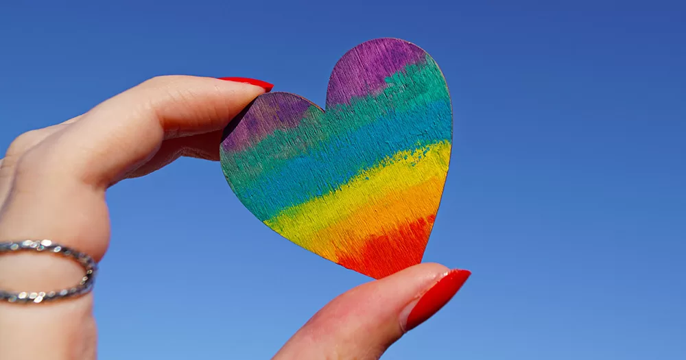 On International Day of Charity we've drawn up a list of 19 of Ireland's top LGBTQ+ charities. The image shows two fingers holding up a rainbow coloured heart against a blue sky.