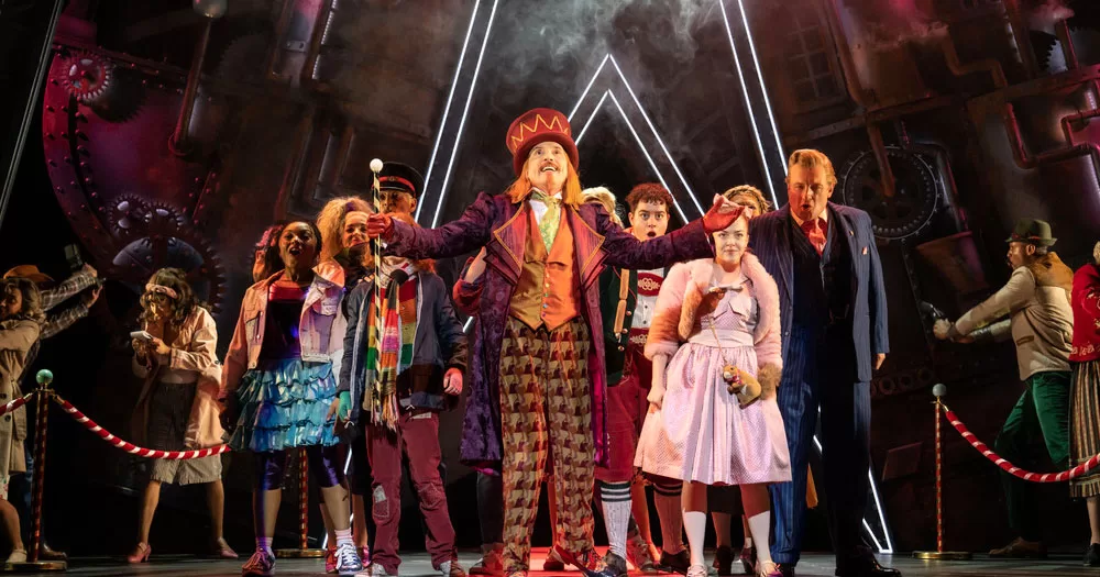 The image shows a scene from Charlie and the Chocolate Factory the Musical. In the photo, the character of Willy Wonka is standing in the centre with a group of people behind him. He is wearing a brown plaid trousers, a brown waistcoat, tails jacket and top hat. He is holding a cane in his right hand and has his arms outstretched looking up.