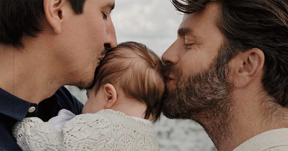 A close up of the cover of Elle Hungary. It shows the side profile of two gay dads, both kissing their baby daughter on the head.