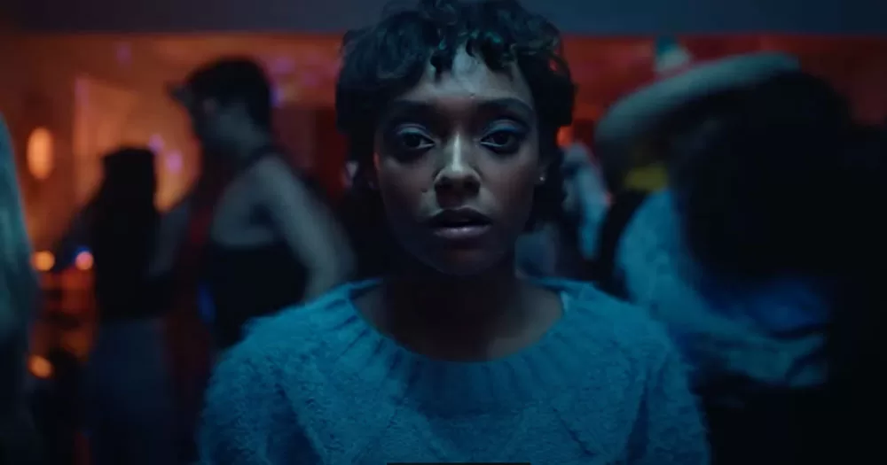The image shows a screenshot from the trailer for Everything Now. It shows the main character Mia at a house party. She is staring directly into the camera and everyone around her is in soft focus. She is wearing a light blue woollen jumper.