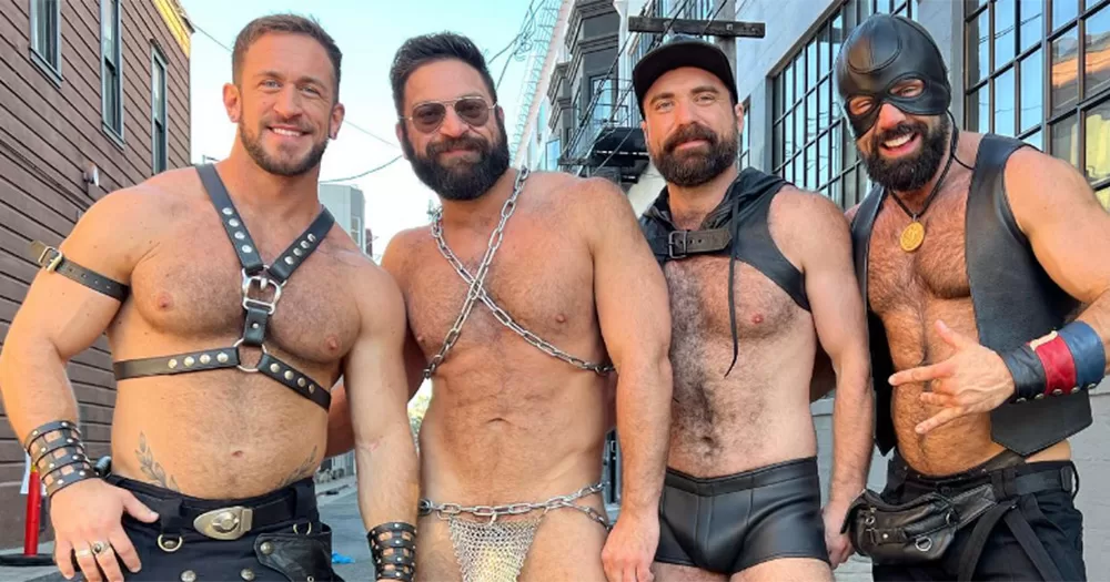 Four people posing for a photo at Folsom 2023. Each person photographed is smiling and shirtless and wearing some form of kink gear. They are photographed from the waist up.