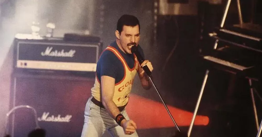 The image shows part of the legacy of Freddie Mercury. It is a photograph of him performing at a concert. He is holding a microphone in one hand and looking down to the audience with other arm outstretched. He is wearing white jeans and a yellow singlet over a navy coloured tshirt.