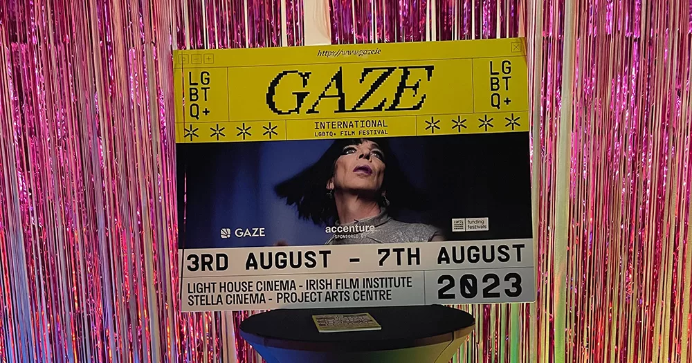 A banners for GAZE 2023. It has black font on yellow and white backgrounds, with a photo of David Hoyle in the middle. It is propped on a table in front of a pink tinsel wall.