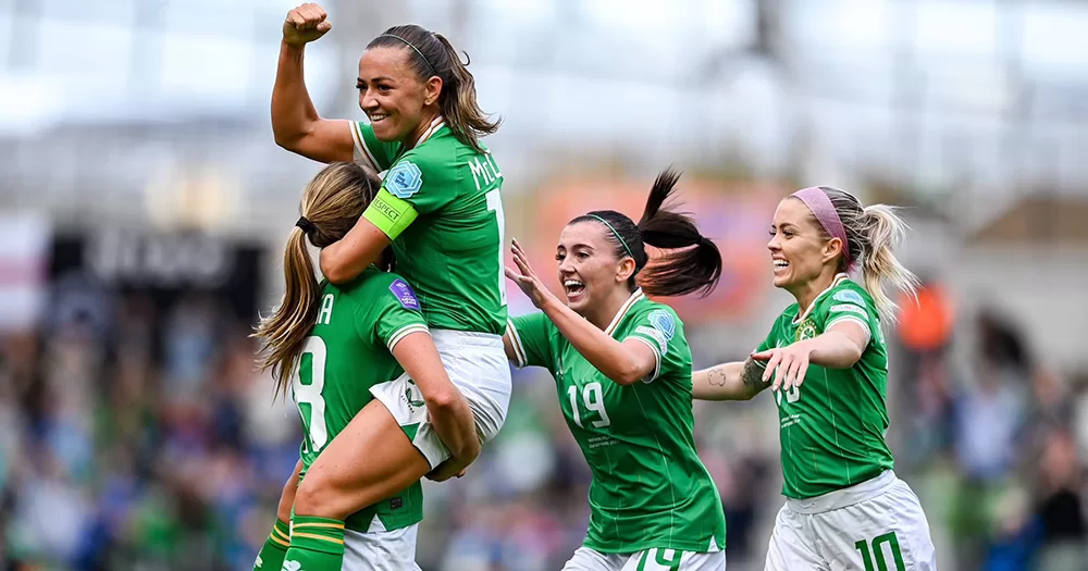 Republic of Ireland players celebrating Kyra Carusa's goal at the Aviva Stadium. In the photo you can see four players, including Carusa, McCabe, Larkin and O'Sullivan. McCabe is being lifted by Carusa, and has her arm raised, while Larkin and Denise are running towards the pair smiling.