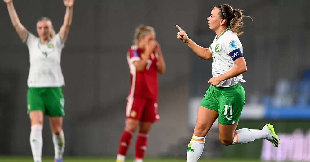 Ireland captain Katie McCabe celebrating her goal against Hungary. The player is seen on the right hand side of the image, running and holding up her right pointer finger in celebration. Blurred in the background we see one Ireland player holding her two hands up in the sky, and one Hungarian player with their hands on their face.