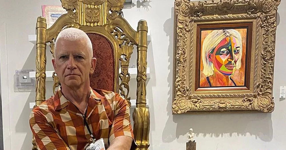 The image shows spoken word artist Maurice Cassidy, who will perform in Outhouse, seated beside a painting titled 'Searching for Secret'. Maurice is sitting in a thrown style seat with gold guilting and ornate carving. Along side the chair is a painting in a regal style picture frame. The frame is also gold guilted with ornate carving. Maurice is wearing a brown and beige gingham shirt which matches the colours of the painting. The painting is an abstract image of a woman with blonde hair and orange skin.