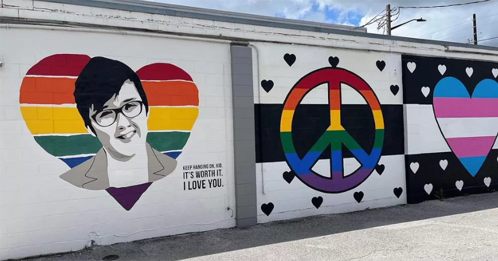 The Lyra McKee mural in Orlando, Florida. It shows a black and white image of Lyra painted on top of a rainbow love heart. It is shown alongside two other LGBTQ+ murals, including one with a rainbow peace symbol and one with a trans love heart.