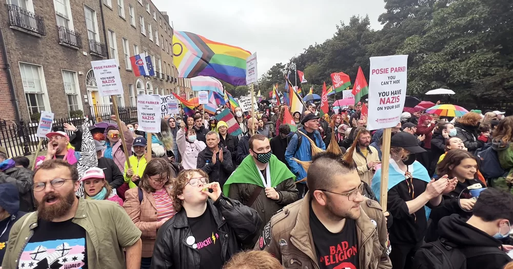 Trans activists protesting in Dublin during a Posie Parker event, with people holding banners and waving Pride and trans flags, shouting and chanting.