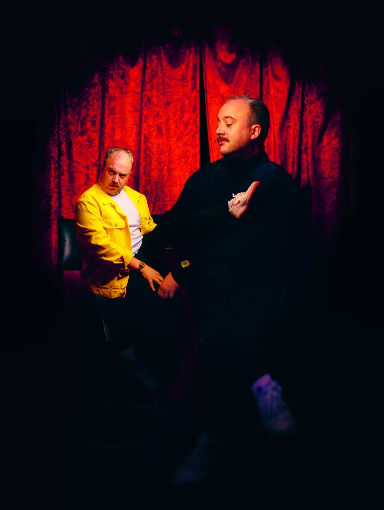 Dublin Fringe 2023 performer Shane Daniel Byrne poses in front of a red curtain holding a puppet of himself.