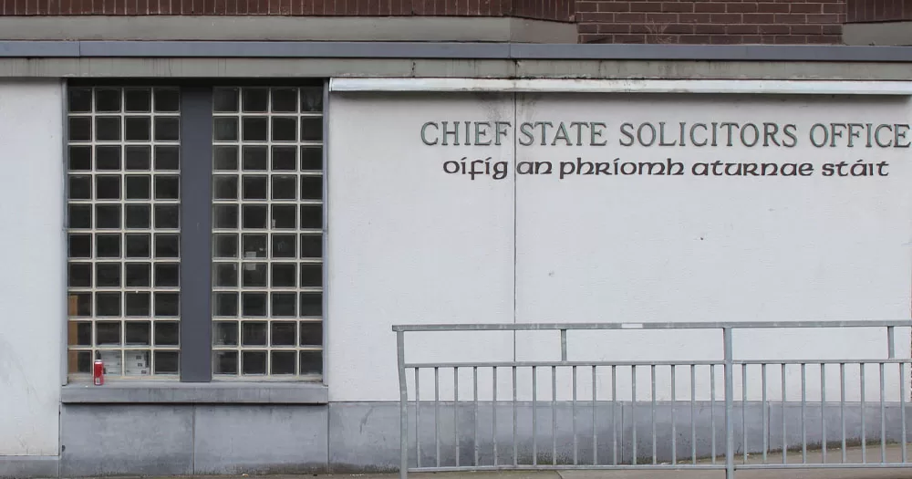 The image shows the front of the Chief State Solicitor's Office in Dublin. It has a grey front with a large window of glass bricks to the left. There is a metal barrier outside at the edge of the footpath closest to the road.